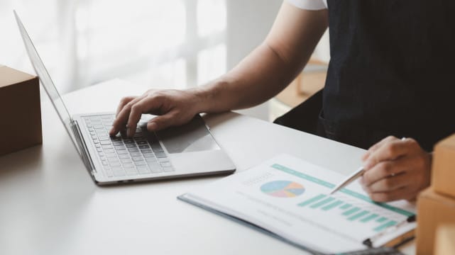 Nesta imagem aparece o braço direito de uma pessoa branca, que está utilizando um notebook e faz anotações no papel