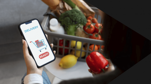 Pessoa segurando celular para fazer compras no mercado. Na imagem, podemos ver uma mão, que segura o celular em um aplicativo de e-commerce de mercado, equanto olha para sua cesta de frutas e verduras.
