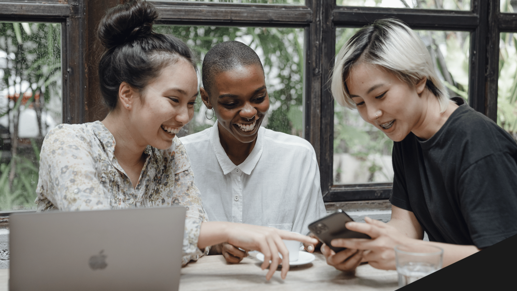 Três pessoas olhando um celular, com diversos ícones de canais/redes sociais em volta para representar o encontro entre a marca e o lead. Opt-in.