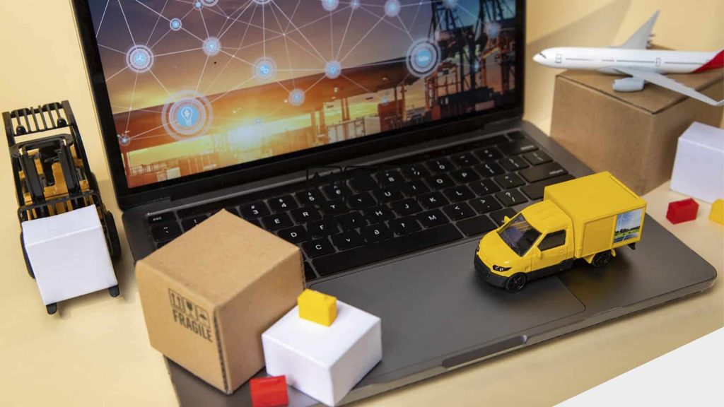 Notebook com uma imagem de vetores conectados na tela, cercado por miniaturas de caixas, caminhões e empilhadeiras, para representar os pedidos.
