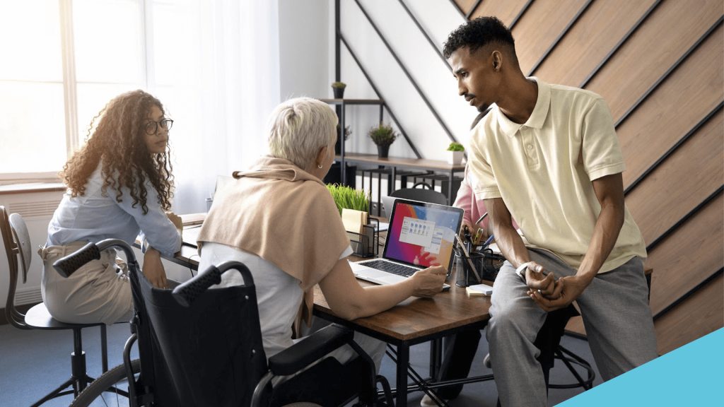 Grupo de pessoas trabalhando numa mesa de escritório. Uma delas está numa cadeira de rodas, em frente a um notebook navegando pela internet.